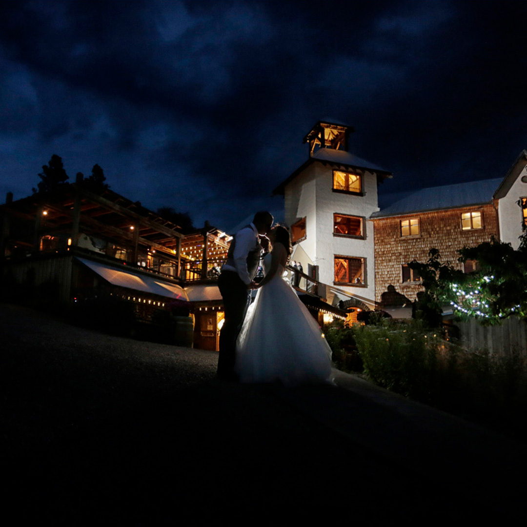 A newly married couple, in their wedding dress and tux, kissing in front of the Hillside Winery venue, the warm lights gently lighting up the husband and wife, with dark blue skies above them. 